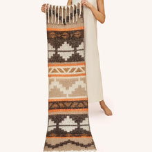 Load image into Gallery viewer, Person wearing a cream-colored dress holding an oversized knit scarf featuring a geometric pattern in black, white, orange, and varying shades of brown, accented with white fringe at the ends, displayed against a white background.