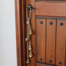 Load image into Gallery viewer, Four gold-colored metal jingle bells wrapped in jute twine, hanging in staggered lengths from a black door handle on a wooden door.