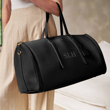Load image into Gallery viewer, A person with cream pants and white top  holding a black weekend bag with the initials  "SLH" on it, standing in front of a white wall.