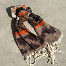 Load image into Gallery viewer, Oversized knit scarf featuring a geometric pattern in black, white, orange, and varying shades of brown, accented with white fringe at the ends. The scarf is tied in a knot and displayed against a gray textured surface.