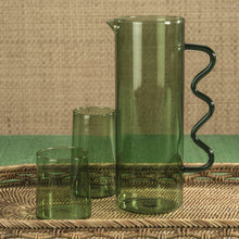 Load image into Gallery viewer, A tall, green glass pitcher with a wavy handle, accompanied by two smaller green glass cups, all placed on a woven mat and textured brown background.