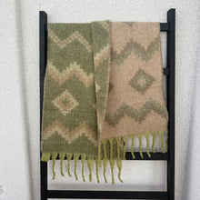 Load image into Gallery viewer, Oversized reversible knit scarf displayed on a black ladder-style rack against a white background. One side features an olive base with cream and tan zigzag and diamond patterns, while the other side has a cream base with olive and brown zigzag and diamond patterns, finished with light green fringe.