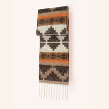 Load image into Gallery viewer, Folded oversized knit scarf featuring a geometric pattern in black, white, orange, and varying shades of brown. The scarf is accented with white fringe at the ends and displayed against a white background.