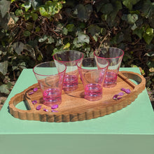 Load image into Gallery viewer, Four pink tinted glasses on a wooden tray sprinkled with purple flowers on top a green table top with bushes in the background.