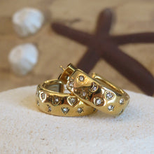 Load image into Gallery viewer, Gold hoop earrings with heart and diamond designs on a sandy surface.