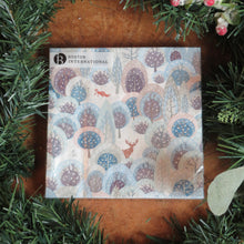 Load image into Gallery viewer, Pack of paper guest size napkins featuring a snowy winter forest design with deer, a fox, and stylized trees in shades of blue, lavender, and gray. Displayed on a wooden surface surrounded by garland and frosted red berries.