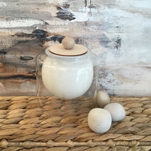 Load image into Gallery viewer, White rounded candle in a glass jar on a wicker stand. Pebbles to the side and an abstract background.