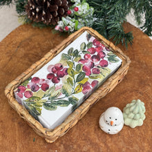 Load image into Gallery viewer, Rectangular rattan napkin holder with a stack of white paper napkins featuring a watercolor-style cranberry and greenery design. Displayed on a wooden surface beside a small white snowman and green tree figurine, with frosted greenery and a pinecone in the background.