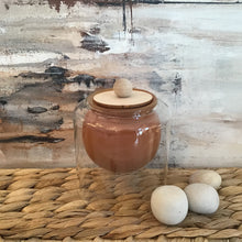 Load image into Gallery viewer, Brown rounded candle in a glass jar on a wicker stand. Pebbles to the side and an abstract background.