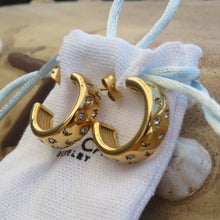Load image into Gallery viewer, Gold hoop earrings with diamond accents on a white jewelry pouch on a sandy background.