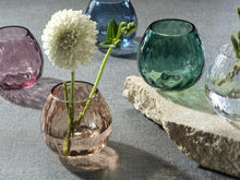 Load image into Gallery viewer, A collection of multipurpose optic vases in various colors including green, blue, clear, brown,  and pink, with a textured design, and a rock. Displayed on a gray textured surface with  flowers in the brown vase.