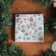 Load image into Gallery viewer, Pack of paper cocktail size napkins featuring a snowy winter forest design with deer, a fox, and stylized trees in shades of blue, lavender, and gray. Displayed on a wooden surface surrounded by garland and frosted red berries.