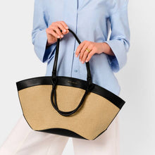 Load image into Gallery viewer, A person wearing a blue shirt and white pants holding a raffia and black tote bag with black handles. On a white background.