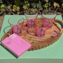 Load image into Gallery viewer, Four pink tinted glasses, gold flamingo cocktail stirs and their  pink packaging box on a wooden tray sprinkled with purple flowers on a green table with bushes in the background.