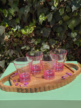 Load image into Gallery viewer, Four pink tinted cocktail glasses on a wooden tray sprinkled with purple flowers, with bushes in the background.