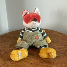 Load image into Gallery viewer, Plush fox with pumpkin orange and white fur, black stitched eyes, a brown nose, yellow hands, and yellow floppy feet. The fox is wearing a green-and-white striped shirt, and moss-colored overalls with a coral patch detail. Fox is sitting upright on a wooden surface, and against a white background.