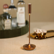 Load image into Gallery viewer, Slim rechargeable table light with a brushed copper finish, featuring a tall narrow cylindrical shade on a thin stem and rounded weighted base, illuminated on a beige surface with a softly blurred beige background featuring a gold tray and bottles.