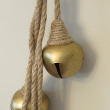 Load image into Gallery viewer, Close-up of a gold-colored metal jingle bell wrapped in and hanging from jute twine against a cream background.