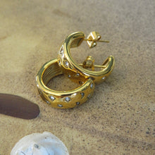 Load image into Gallery viewer, Gold hoop earrings with embedded  diamond stones on a sandy surface.