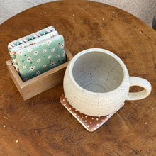 Load image into Gallery viewer, A cream-colored mug with black speckles sitting on a red square shaped resin coaster with white polka dots, next to a wooden box with three square shaped resin coasters with various holiday designs. One coaster is light green with green polka dots and white flowers with a red center. One coaster is white with green and red various candies. One coaster has green, red, and white diagonal stripes. White polka dots on the green stripes.  On a wooden surface with a partial view of a white textured background.