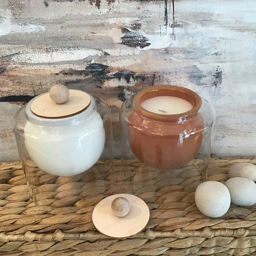 White and brown rounded candles in glass jars on a wicker stand. Pebbles to the side and an abstract background. 