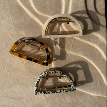 Load image into Gallery viewer, Three half-moon metal claw clips in ivory, tortoiseshell, and leopard patterns displayed on a beige textured surface with  soft curved light reflections.