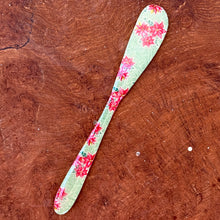 Load image into Gallery viewer, A teal canapé knife with a pink floral design. On a brown wooden surface.