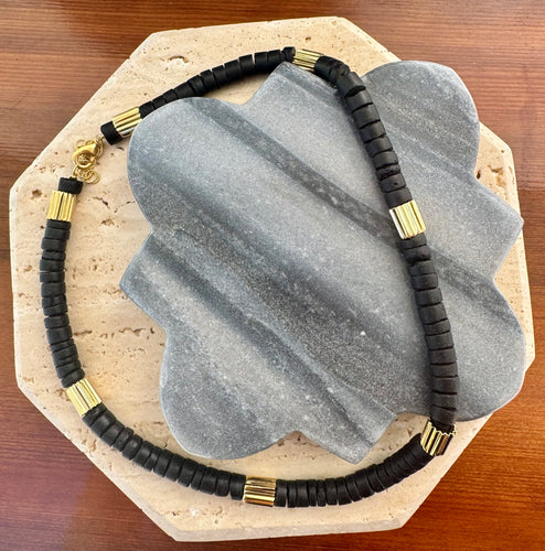 Black disc-bead necklace accented with multiple ribbed gold tube beads, displayed in a loose circle on a quatrefoil-shaped gray stone tile layered over a beige travertine slab on a wooden surface. 