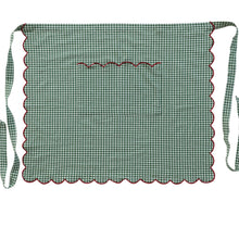 Load image into Gallery viewer, Holiday Apron, green cotton gingham waist apron displayed flat with red scalloped trim along the edges and bib pocket, with gingham waist ties extending at the sides, against a cream background. 