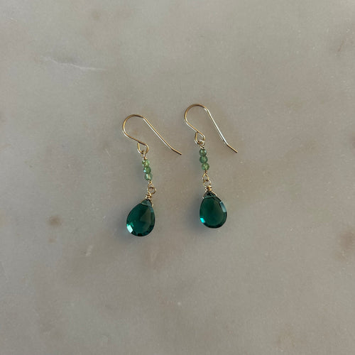 A pair of gold drop earrings with three small green beads down to a green teardrop gem. White marble background. 