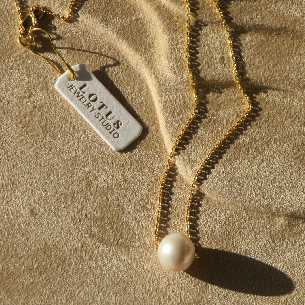Gold chain necklace with a single round white pearl pendant, displayed on a textured tan surface with a white brand tag that reads 