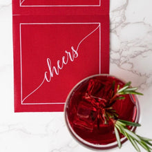Load image into Gallery viewer, Red cotton cocktail napkin with a white border and "cheers" in white script, displayed on a white marble surface beside a red cocktail garnished with rosemary. 