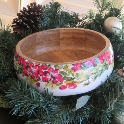 Round mango wood bowl with a white enamel exterior decorated with a cranberry wreath design of red berries and green leaves, displayed nestled in green garland with pinecones and silver ball ornaments. 