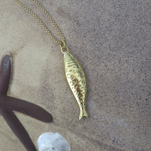 Load image into Gallery viewer, Gold fish-shaped pendant on a chain with a starfish and shell on a sandy surface