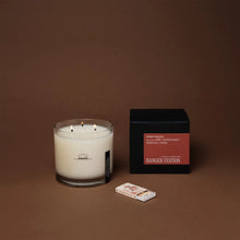 Load image into Gallery viewer, Ranger Station Cowboy Christmas candle with lit triple wicks in a clear glass jar featuring a black cowboy illustration, displayed beside its black box with a red label reading &quot;amber, Montana juniper, frankincense, whiskey,&quot; and a small matchbox on a warm brown surface. 