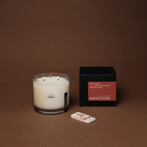 Ranger Station Cowboy Christmas candle with lit triple wicks in a clear glass jar featuring a black cowboy illustration, displayed beside its black box with a red label reading 