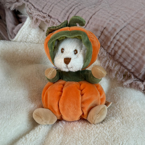 White plush bear with cream-colored hands and feet, dressed in a soft orange pumpkin costume with a green center and a green-and-orange hood featuring a green stem and leaf detail. The bear is sitting on a plush white blanket with a mauve fringed textile in the background.  