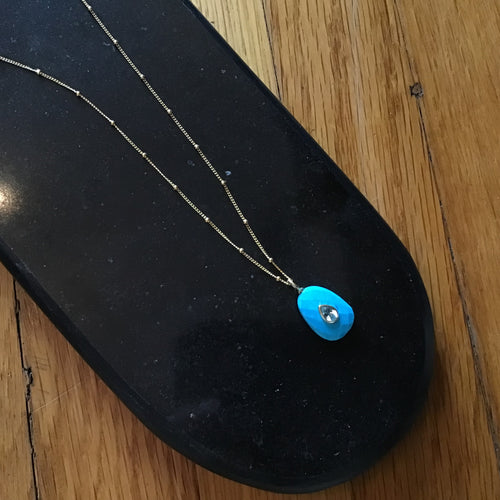A gold chain necklace with a cut turquoise pendant and aquamarine teardrop on a black display on a wooden surface.