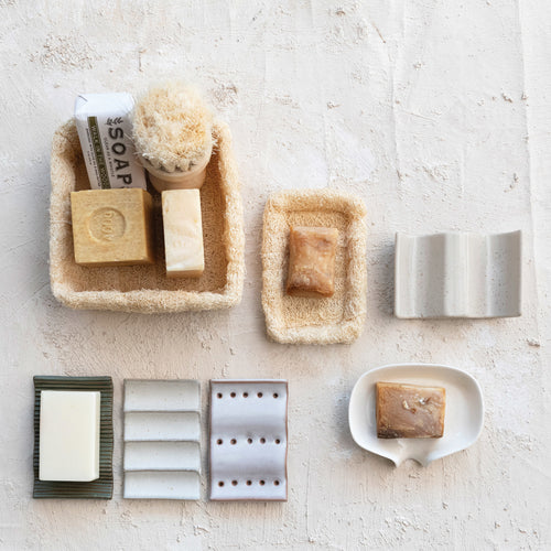 A collection of stoneware soap dishes in various styles displayed alongside soap bars and a loofahs, on a cream -colored textured surface.