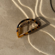 Load image into Gallery viewer, Tortoiseshell half-moon metal claw clip with gold hardware displayed on a beige textured surface with soft curved light reflections. 