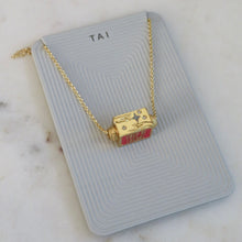 Load image into Gallery viewer, Gold-plated necklace featuring a small rectangular spinner pendant with enamel detailing, star motifs, and engraved  accents, suspended on a gold-tone chain, displayed on a light gray TAI jewelry card on a white marble background.