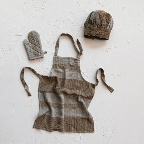 Children's brown and grey cotton apron set with a chef hat and oven mitt laid out on a white surface.
