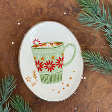 Load image into Gallery viewer, White, oval, ceramic holiday dish with a green mug with a pink flower design. Brown drink , white marshmallows, and a red and white candy cane in mug. Orange dots and designs at the bottom of the dish. On a wooden surface, surrounded by fir tree clippings .