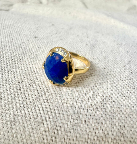 Double-gold-plated brass ring featuring an oval lapis stone set in a prong setting with small CZ accent stones, displayed on a light textured fabric surface.  