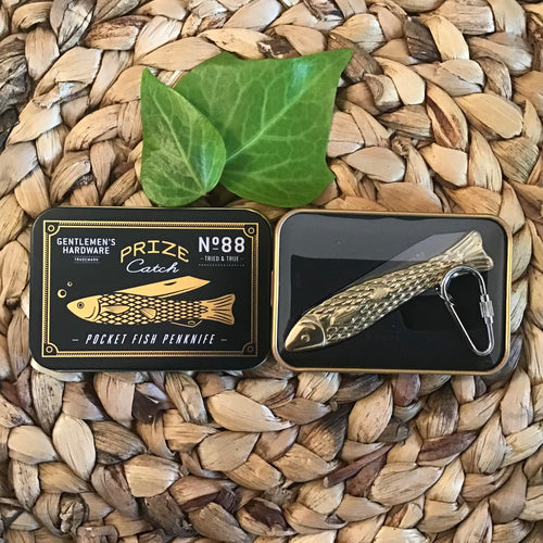 Gold penknife in the shape of a fish attached to a silver carabiner. The penknife is in the bottom of a black and gold tin. The top of the tin sits to the left with the penknife printed on it. All sits next to two leaves on a braided brown placemat. 