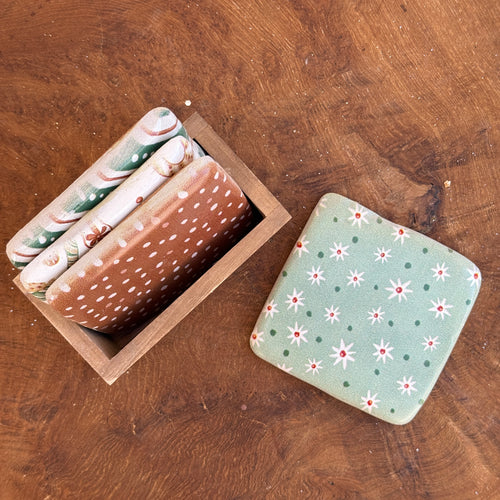 A light green colored square resin coaster with green polka dots and white flowers with a red center, next to a wooden box with three square shaped resin coasters. First coaster is red with white polka dots. Second coaster is white with red and green various candies. Third coaster has red, white, and green diagonal stripes. White polka dots on the green stripes. Coasters sitting on a brown wooden surface. 