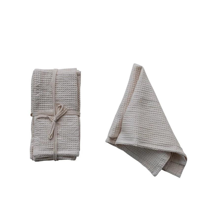A set of folded white cotton waffle napkins ,wrapped with a white  ribbon . Next to an identical white unfolded napkin. On a white background.  