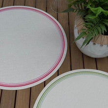Load image into Gallery viewer, Round placemats with striped embroidered edges, one pink and one green . On a wooden surface next to a potted plant.