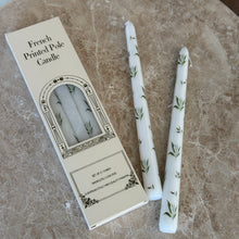 Load image into Gallery viewer, Two white taper candles with green botanical designs on a brown marble surface, next to their packaging. 
