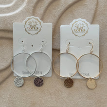 Load image into Gallery viewer, Two pairs of hoop earrings displayed on a LOTUS brand cards, one sterling silver and one 14K gold-filled, each featuring a hand-hammered round disc charm, shown against a tan textured surface. 